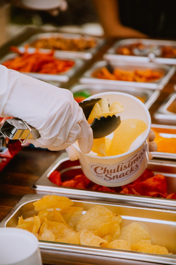 Gloved hand serving potato chips into a cup from a snack bar with various snacks on display.